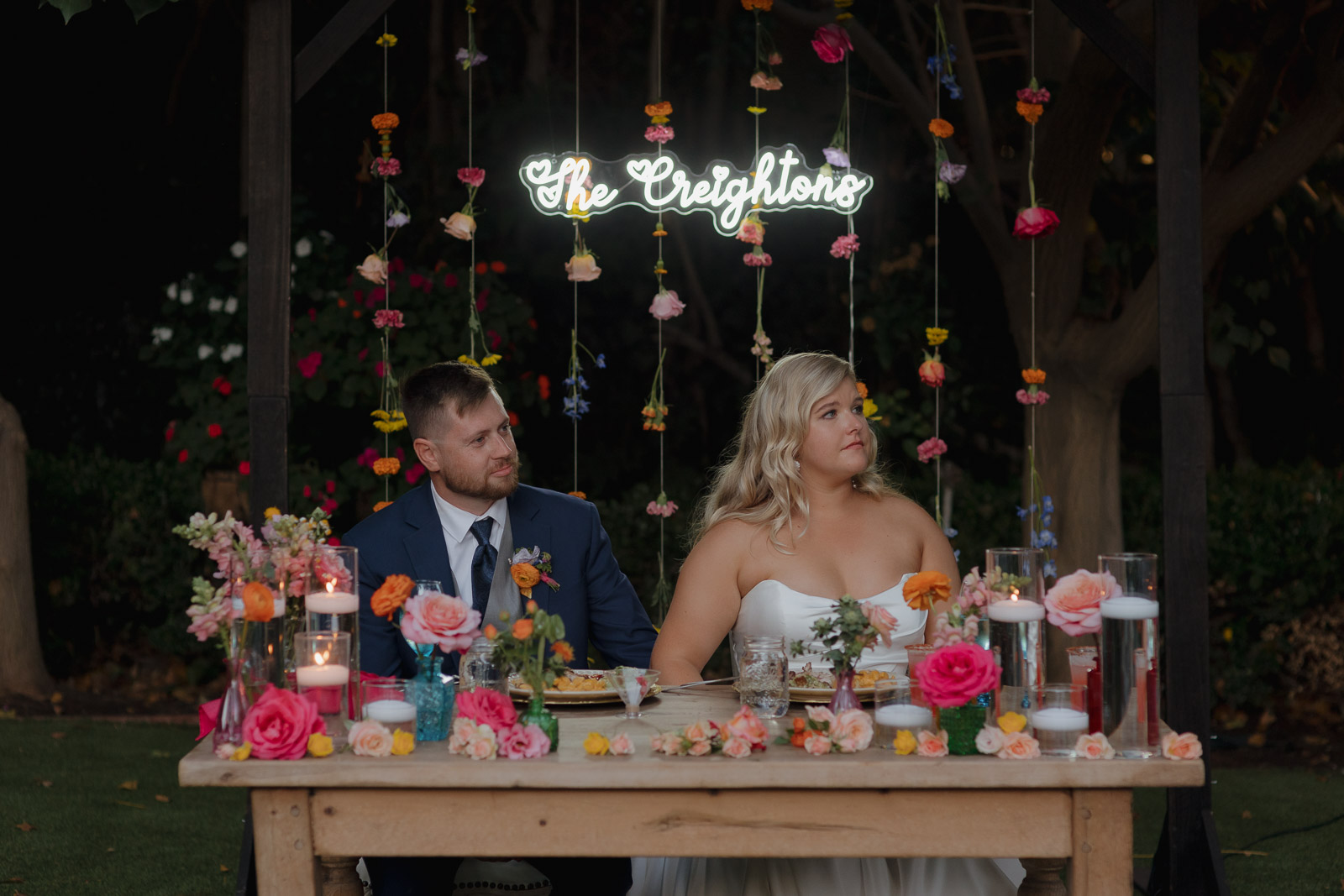 Bride and groom listen to toasts with a wildflower backdrop and neon last name sign